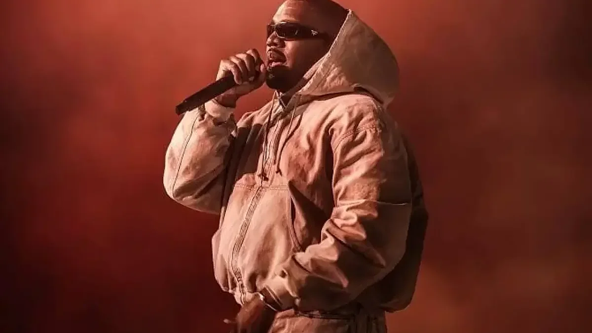 Ye tampil di panggung konser dengan asap merah tebal dan cahaya panggung ungu, mengenakan jaket cokelat, kemeja putih, dan kacamata hitam